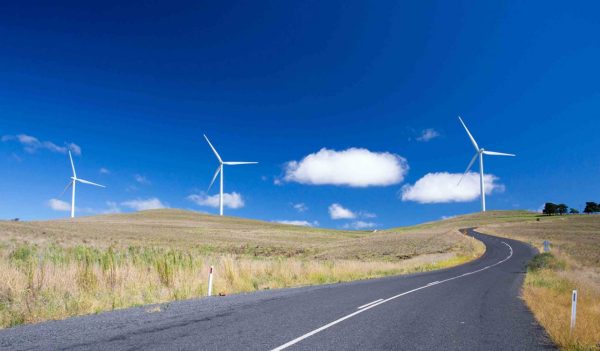 Narrow road surrounded by fields with three wind turbines in the distance against a blue sky.