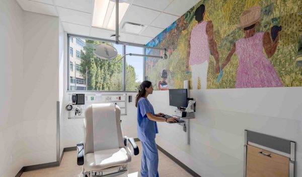 A patient room with a hospital worker looking at a computer. A mural of a woman and girl in a field fills the wall. Image credit: Kyle Jeffers